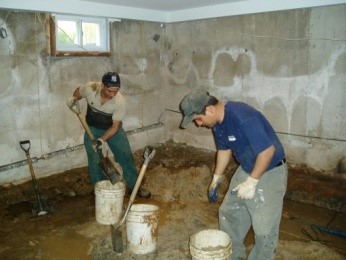 Two workers excavating a basement with shovels and buckets, removing soil and debris to prepare for waterproofing, showcasing A.M. Shield Waterproofing's basement excavation services.