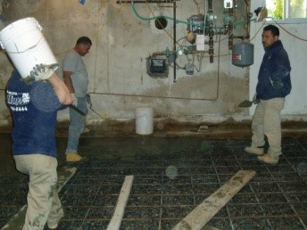 Workers installing a concrete slab in a basement, with visible plumbing and wall moisture issues, illustrating A.M. Shield Waterproofing's foundation repair services.