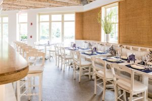 Interior of Kyma Restaurant in North Hills, featuring white wooden tables and chairs, neatly set with blue napkins and glassware, showcasing a bright and inviting dining atmosphere.