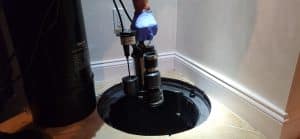 A person wearing a blue glove adjusts a sump pump inside a black basin on a tiled floor. The setting is indoors, with a white wall in the background.