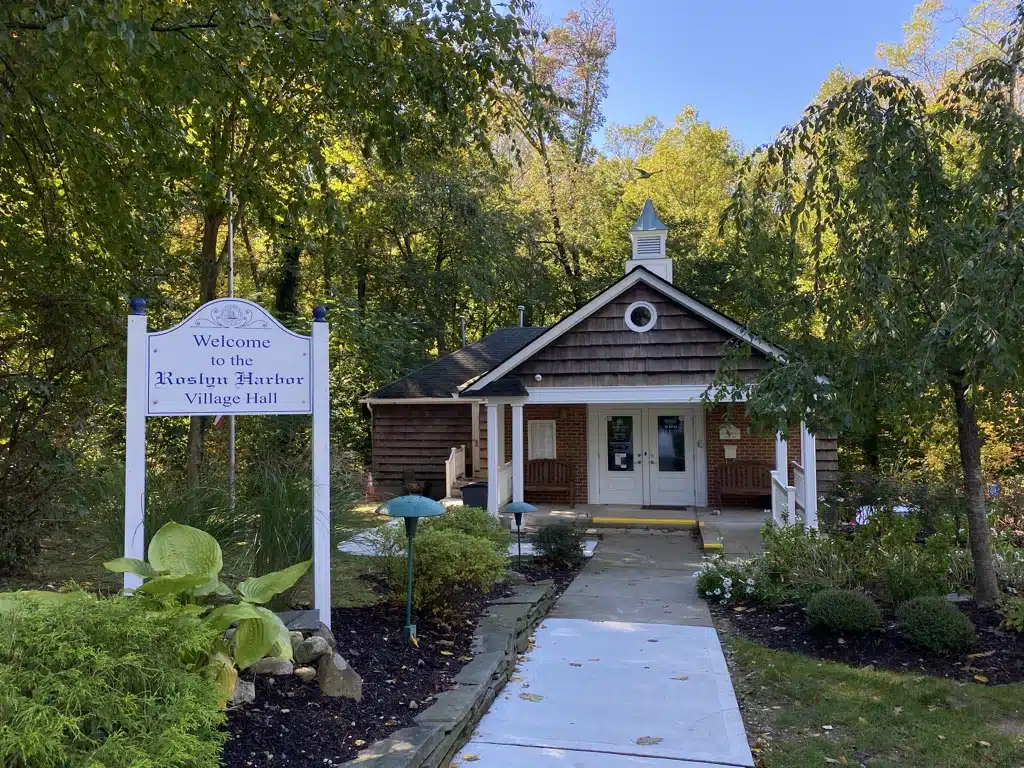 Welcome sign for Roslyn Harbor Village Hall in a landscaped setting, featuring a charming wooden building surrounded by greenery and pathways.