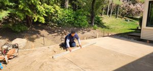 A person kneels on packed dirt, smoothing a surface with a long wooden tool. Nearby are trees and a house. The scene conveys a sense of industriousness and tranquility.