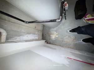 Dimly lit corner of a basement with peeling paint and rusted pipes. A person in white sneakers stands on a cracked floor, conveying neglect.