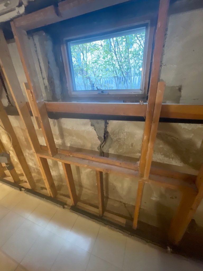Basement wall with exposed wooden framing and a small window showing trees outside. Visible crack in concrete suggests wear, evoking a neglected ambiance.